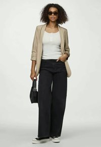 Beige Blazer über einem weißen Oberteil mit Knöpfen, schwarze weit geschnittene Jeans, schwarze Sneakers und schwarze Handtasche. Person mit dunklen Sonnenbrillen und lockigem Haar.