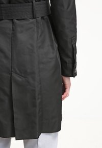 Vue de dos d’une personne portant un manteau noir ceinturé avec des poignets boutonnés et un pantalon de couleur claire.