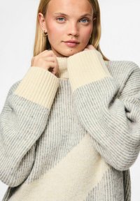 Beež ja hall triibuline kampsun kõrge kraega ning soonikute äärtega, millel on pehme tekstuur ja diagonaalne muster üle keha.