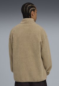 Chaqueta de forro polar beige con cuello alto, corte oversize, superficie texturizada y hombros caídos, mostrada desde la parte trasera.
