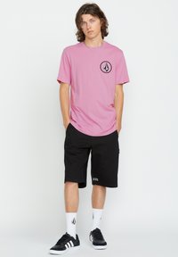 Pink cotton t-shirt with a black logo on the chest, paired with black shorts, white socks, and black sneakers with white stripes.