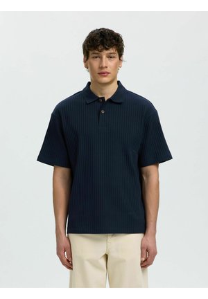 Giovane uomo con capelli ricci che indossa una polo a maniche corte a coste blu navy e pantaloni beige, in piedi davanti a uno sfondo chiaro uniforme.