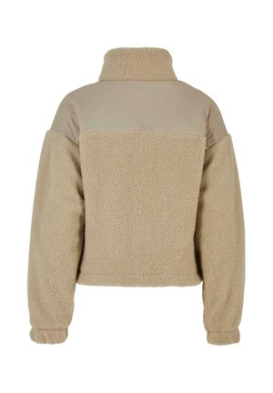 Smėlio spalvos teddy fleece striukė su aukštu apykakle ir lygiu audinio skydu virš apatinės nugaros ir pečių, ilgi rankogaliai su elastingomis rankogalių juostelėmis.