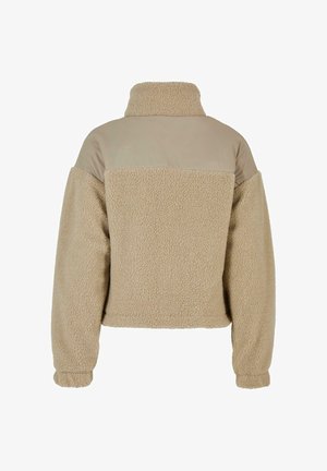 Smėlio spalvos teddy fleece striukė su aukštu apykakle ir lygiu audinio skydu virš apatinės nugaros ir pečių, ilgi rankogaliai su elastingomis rankogalių juostelėmis.