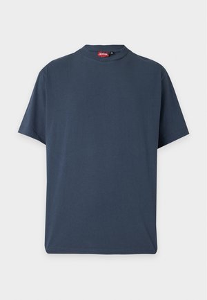 Mand stående iført en mørkeblå t-shirt, sorte knælange shorts, hvide sokker og hvide sneakers på en ensfarvet lys baggrund.