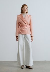 Ροζ σακάκι με ζώνη και διπλό κούμπωμα, συνδυασμένο με λευκό παντελόνι φαρδιάς γραμμής και μπεζ παπούτσια με τακούνι. Ομαλές υφές και ραμμένη εφαρμογή.