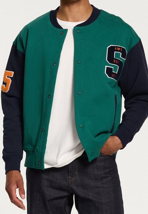 Chaqueta de béisbol verde y azul marino con cuello redondo, cierres frontales a presión, detalles bordados y un gran logo "S" en el pecho.
