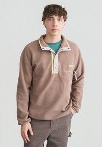 Νεαρός με σγουρά μαλλιά που φορεί καφέ φλις μπλούζα με κουμπιά στον γιακά και τσέπη με φερμουάρ στο στήθος, ποζάροντας με το ένα χέρι στην τσέπη.