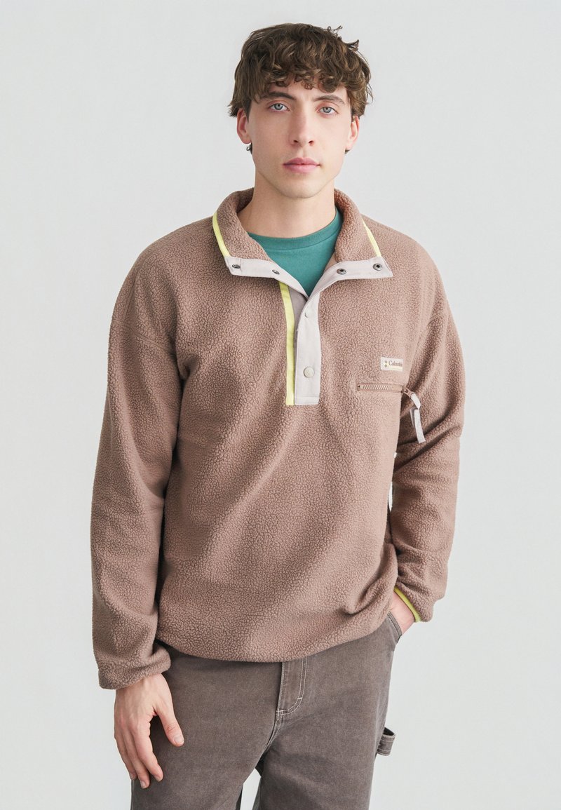 Νεαρός με σγουρά μαλλιά που φορεί καφέ φλις μπλούζα με κουμπιά στον γιακά και τσέπη με φερμουάρ στο στήθος, ποζάροντας με το ένα χέρι στην τσέπη.