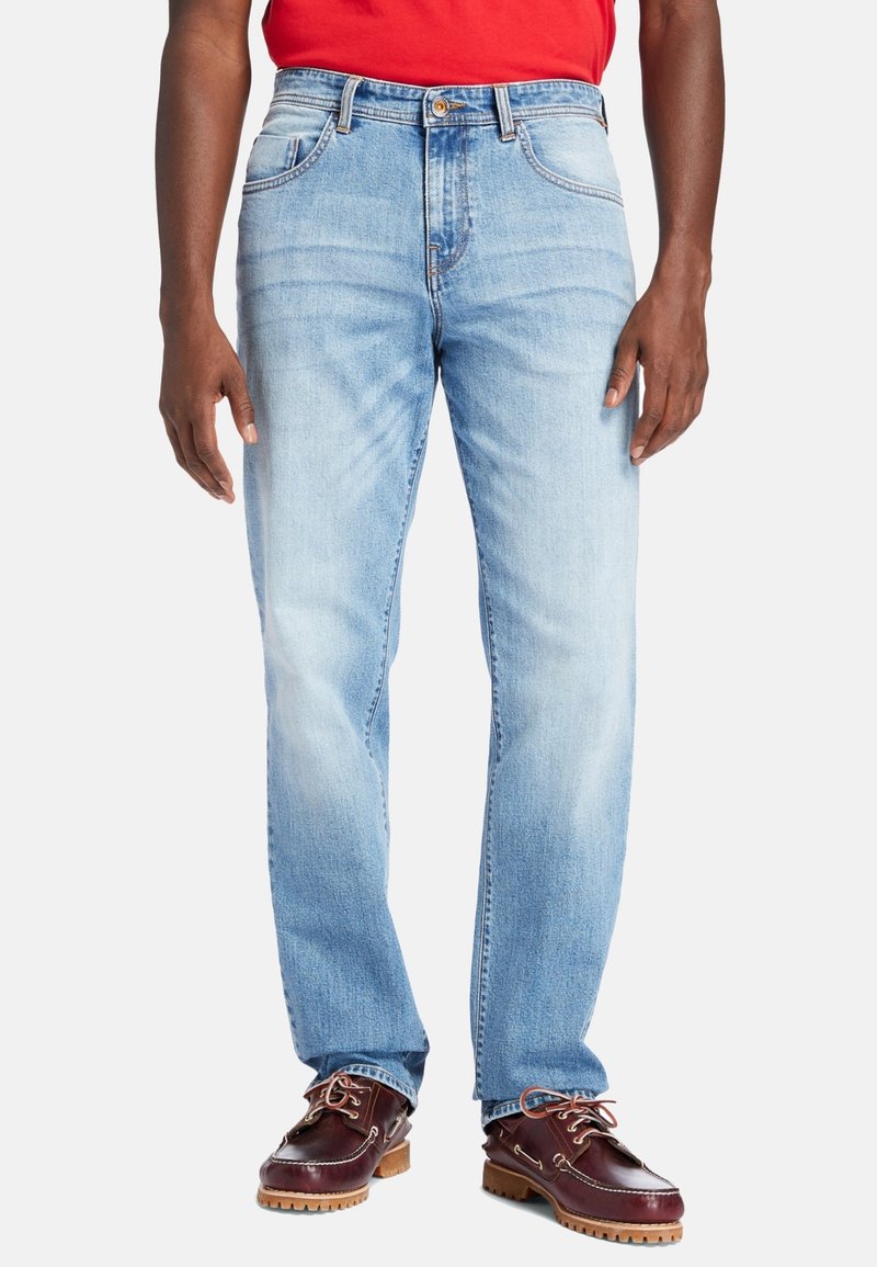 Personne portant un jean droit bleu clair, des chaussures bateau en cuir marron et une chemise rouge, debout sur un fond blanc uni.