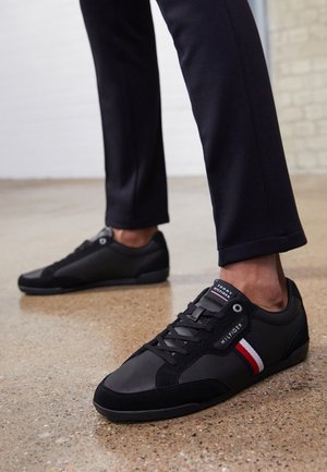 Persona con zapatillas negras Tommy Hilfiger con rayas rojas, blancas y azules, combinadas con pantalones oscuros sobre un suelo de hormigón pulido.