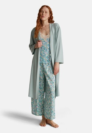 Femme aux longs cheveux roux, portant un ensemble de pyjama à fleurs bleu clair et un peignoir assorti bleu clair, pieds nus, se tenant contre un fond blanc.