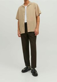 Uomo che indossa una camicia beige a maniche corte con bottoni, una maglietta bianca sotto, pantaloni marroni scuri e mocassini neri, in piedi contro uno sfondo neutro.