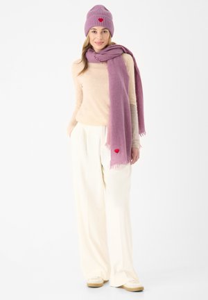 Lila Strickmütze mit einem roten Herz, passender lila Schal, beiger Pullover und weißen weiten Hosen, kombiniert mit hellen Schuhen.