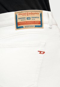 Λευκά τζιν από denim με δερμάτινη ετικέτα Diesel Industry σε χρώμα ταμπά και κόκκινο κεντητό λογότυπο στην πίσω τσέπη, συνδυασμένα με μαύρο πάνω μέρος.