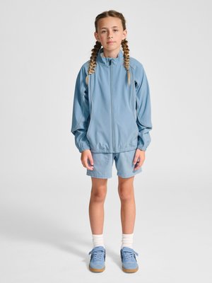 Junges Mädchen mit geflochtenem Haar steht in einer hellblauen Jacke mit Reißverschluss, passenden Shorts, weißen Socken und blauen Sneakers vor einfachem Hintergrund.