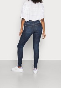 Vaqueros ajustados de color azul oscuro con dos bolsillos traseros, combinados con una camisa suelta de patrón a cuadros en blanco y zapatillas blancas.