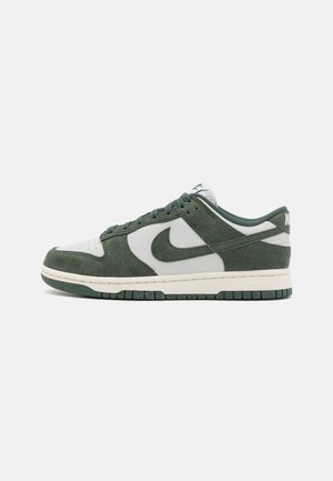 Zapatilla baja en ante verde y cuero blanco con el logo de Nike, diseño con cordones y suela moteada verde oscuro sobre fondo blanco.