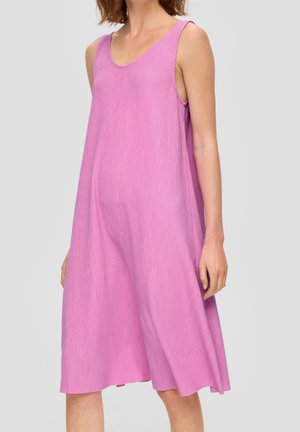 Mouwloze losse roze jurk tot op de knie gedragen door een persoon met schouderlang bruin haar, staande tegen een eenvoudige achtergrond.