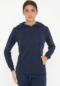 Felpa con zip di colore navy, dotata di cappuccio con coulisse, polsini e orlo a costine, caratterizzata da una texture di tessuto liscia e tasche laterali.