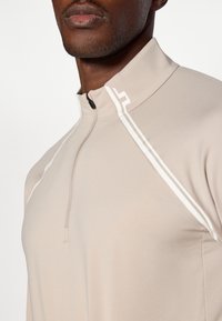 Jasbeżowy sweter typu quarter-zip wykonany z gładkiego materiału, z wysokim kołnierzem i białymi akcentami na ramionach.