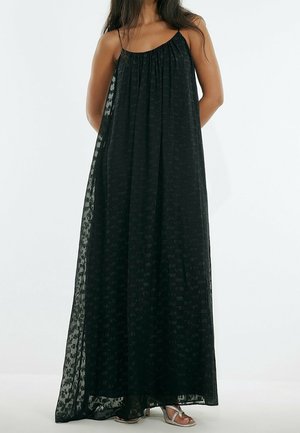 Vrouw draagt een lange, zwarte, mouwloze jurk met textuur en dunne bandjes, gecombineerd met zilveren open-toe hakken, staand tegen een effen achtergrond.