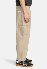 Beige cargo bukser med sidelommer, lavet af let stof. Lige pasform med en afslappet fit, parret med sorte sneakers.