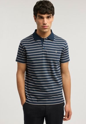 Giovane uomo che indossa una polo blu navy con strisce orizzontali bianche, in piedi con le mani in tasca davanti a uno sfondo neutro.