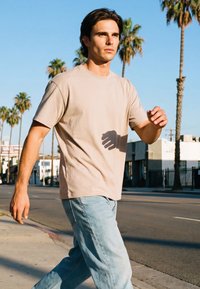 Mladi moški v bež majici in svetlo modrih kavbojkah hodi po sončni mestni ulici s palmami in stavbami v ozadju.