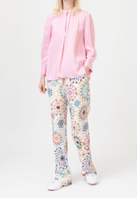 Femme portant un blouse à manches longues rose, un pantalon à motifs multicolores avec des designs circulaires, et des baskets blanches, se tenant devant un fond uni.