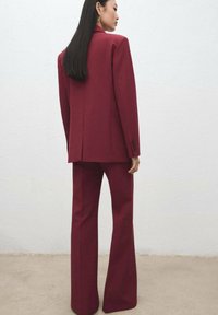 Femme aux longs cheveux noirs, portant un blazer bordeaux ajusté et un pantalon évasé assorti, debout devant un mur clair uni.