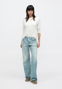 Γυναίκα που στέκεται φορώντας λευκό πουκάμισο με μακριά μανίκια, ανοιχτό μπλε τζιν μπόξερ και πράσινα αθλητικά παπούτσια σε λευκό, απλό φόντο.