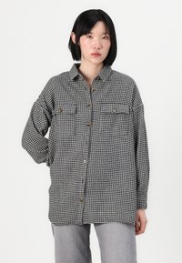 Oversized black and white houndstooth shirt with front buttons, two chest pockets, and a collared neckline. Made of textured fabric.