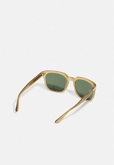 Óculos de sol quadrados com uma armação translúcida castanho claro, lentes verdes e hastes finas, apresentando detalhes em prata nas dobradiças.