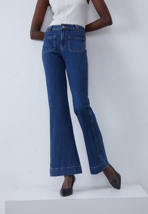 Kvinde iført højtaljede blå flare jeans, hvid ærmeløs top og sorte spidse hæle står op ad en hvid væg.