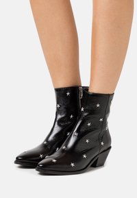 Botas de piel negras hasta el medio de la pantorrilla con tachuelas en forma de estrella plateadas, cremalleras laterales, punteras puntiagudas y tacones cuadrados, llevadas sobre piernas desnudas.