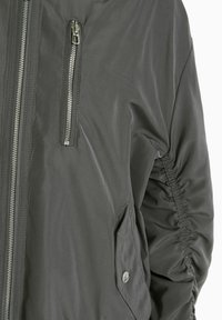 Šedá bombera s zipovou kapsou na hrudi, postranní kapsou a nařasenými detaily na rukávech. Vyrobena z hladké, odolné tkaniny.