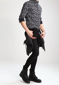 Camisa negra de botones con patrones abstractos en blanco, combinada con jeans ajustados negros y botas negras de cordones. Una chaqueta está atada a la cintura.