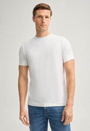 Uomo che indossa una maglietta bianca semplice a maniche corte e jeans blu, in piedi davanti a uno sfondo chiaro.