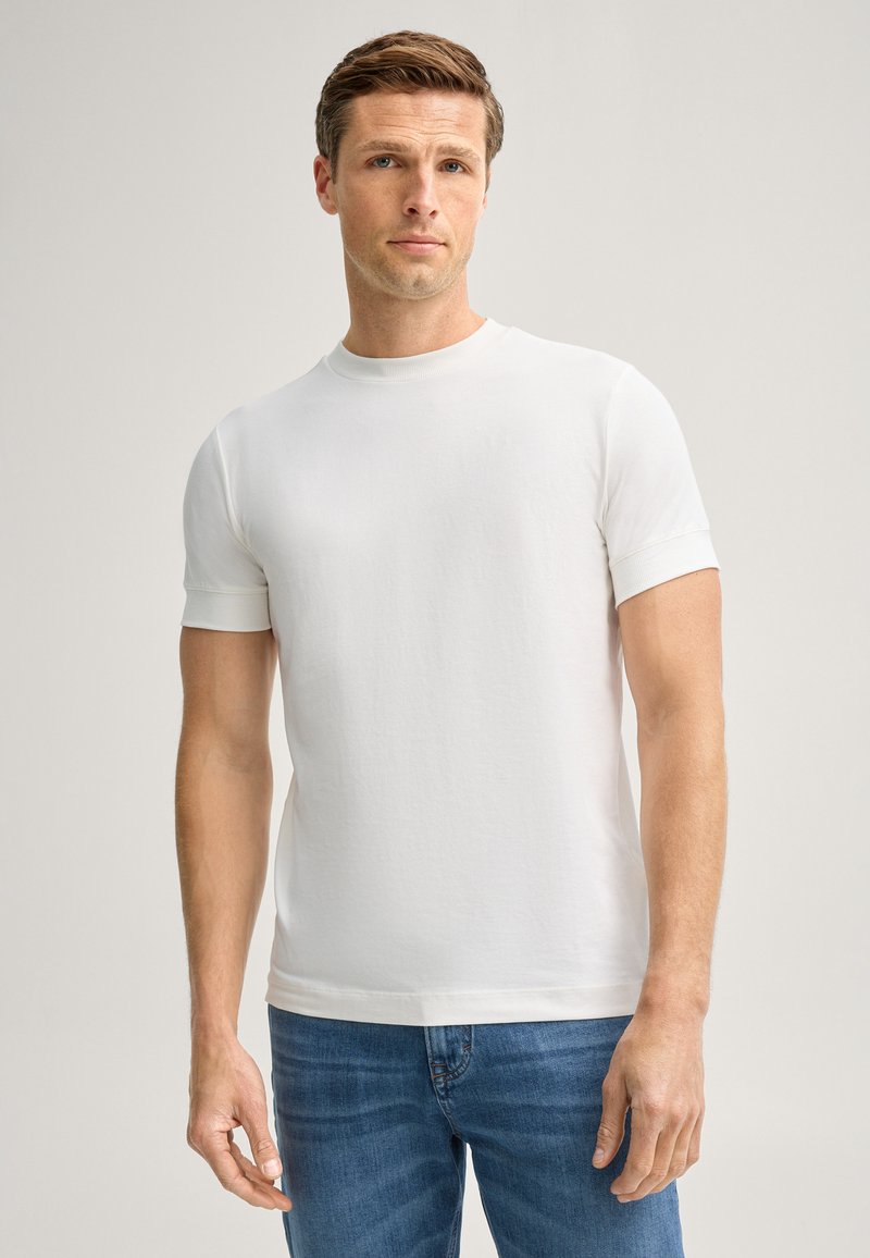 Mann trägt ein schlichtes weißes Kurzarm-T-Shirt und blaue Jeans und steht vor einem hellen Hintergrund.