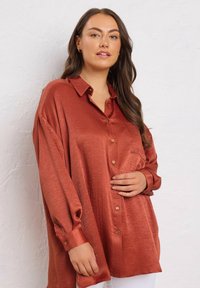 Woman with long brown hair wearing a loose, shiny rust-colored button-up shirt, standing against a white textured wall.