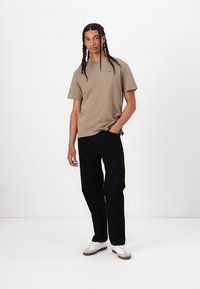 Khaki bomulds T-shirt med korte ærmer og et lille logo på brystet, parret med sorte bukser og hvide sneakers. Ensartet hvid baggrund.