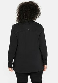 Person mit lockigem Haar, der eine schwarze Jacke mit langen Ärmeln trägt, von hinten vor einem schlichten weißen Hintergrund gezeigt.