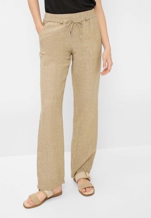 Vrouw draagt beige linnen broek met trekkoord en zijzakken en beige open-toe platte sandalen tegen witte achtergrond.