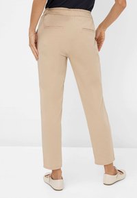 Pantalon beige à coupe décontractée, taille élastique et deux poches arrière. Le modèle porte des ballerines de couleur claire. Texture simple et lisse.