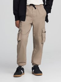 Beżowe spodnie cargo z elastycznym pasem, troczkiem, bocznymi kieszeniami oraz ściągaczami przy nogawkach; wykonane z trwałej tkaniny bawełnianej mieszanej.