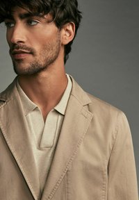 Blazer beige avec un polo crème texturé en dessous. Le blazer possède des revers à cran et des détails de couture visibles. Cheveux bien coiffés et barbe de trois jours.