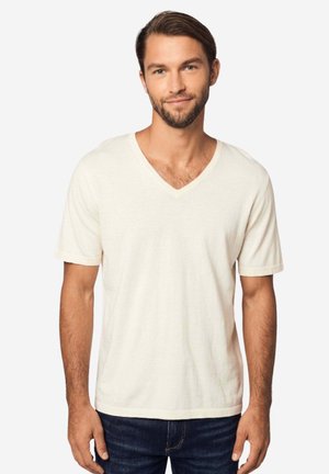 Mann mit kurzen braunen Haaren und Bart, trägt ein schlichtes cremeweißes T-Shirt mit V-Ausschnitt und kurzen Ärmeln sowie dunkelblaue Jeans, steht vor weißem Hintergrund.