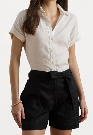Femme portant une chemise blanche à manches courtes boutonnée rentrée dans un short noir taille haute avec une ceinture nouée et un bracelet en or au poignet gauche.