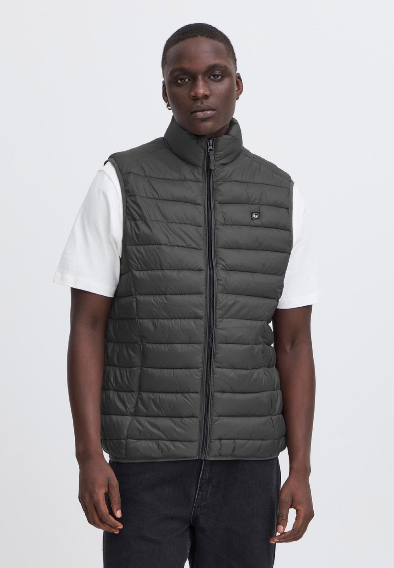 Jeune homme portant une veste matelassée sans manches gris foncé avec une fermeture éclair devant, par-dessus une chemise blanche à manches courtes et un pantalon foncé.
